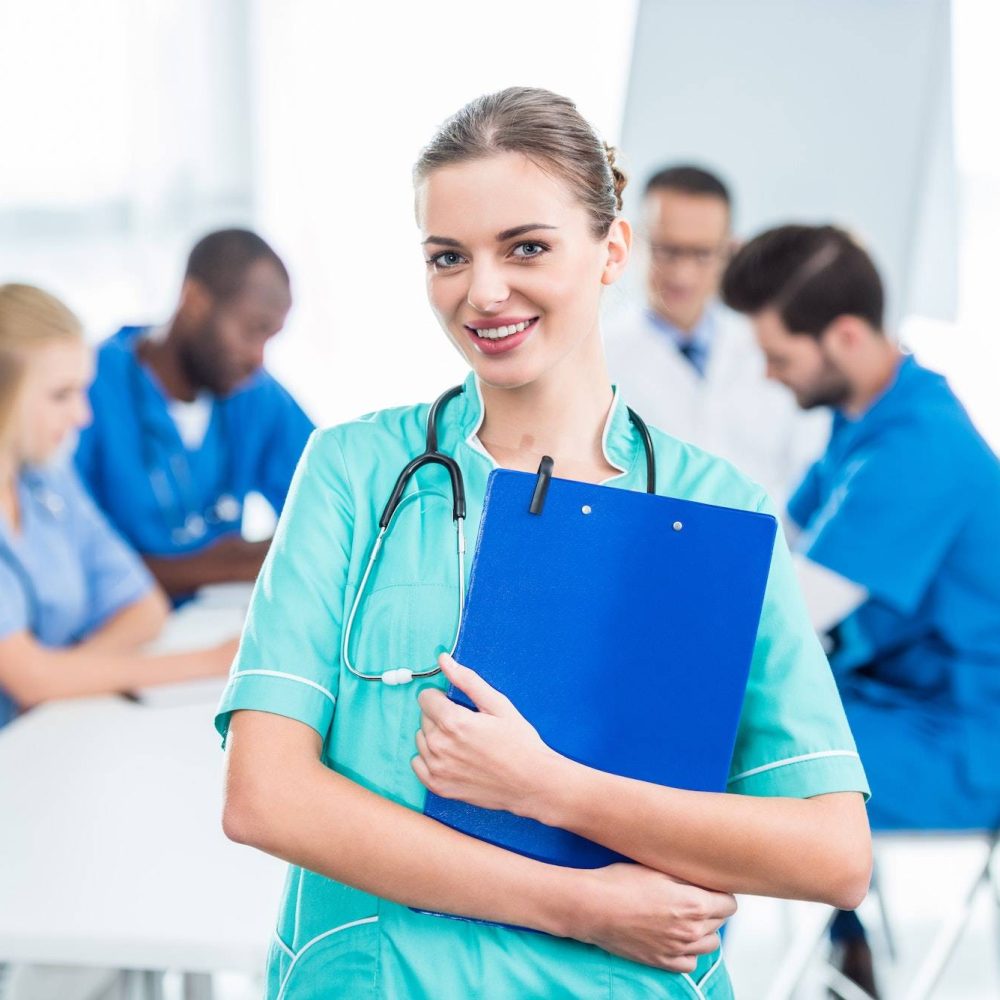 attractive young nurse holding clipboard with colleagues sitting at table on background